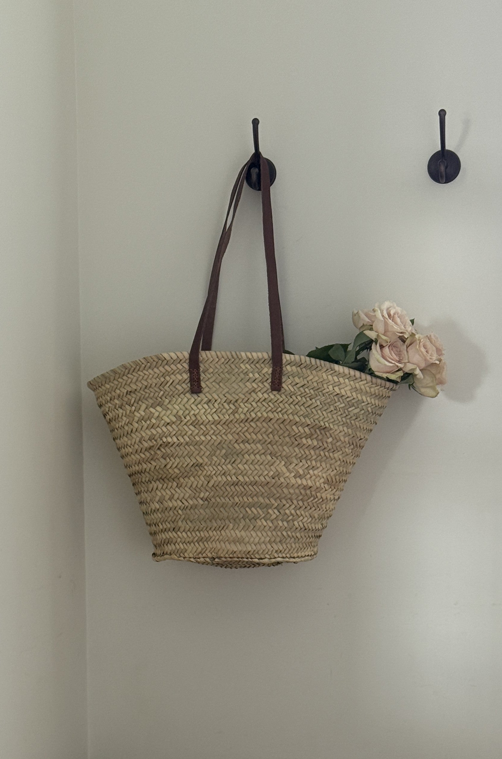 Woven bag with brown handles hanging on a wall hook next to a small bouquet of flowers.