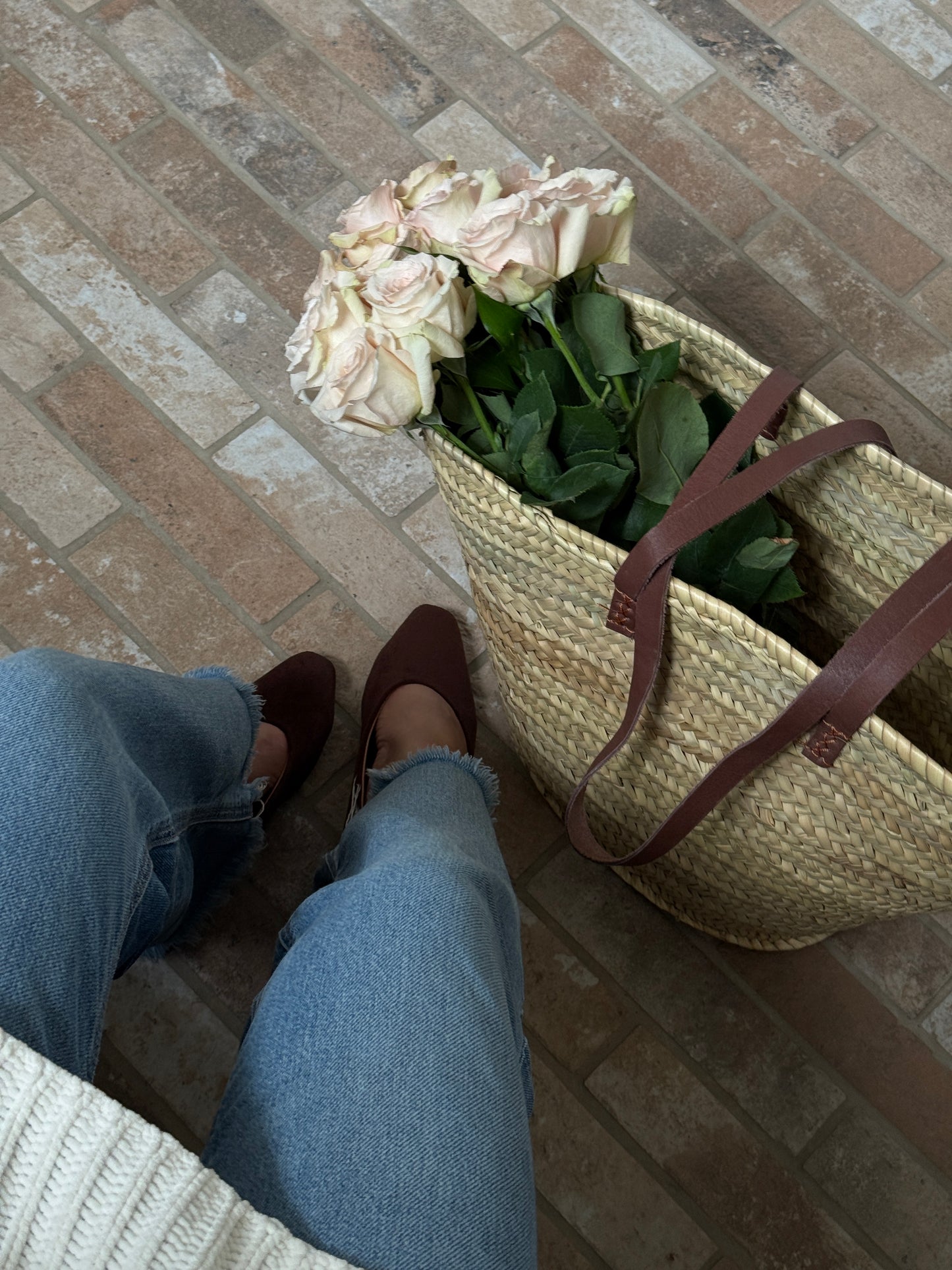 Person wearing jeans and brown shoes standing next to a woven basket with flowers on a brick floor.